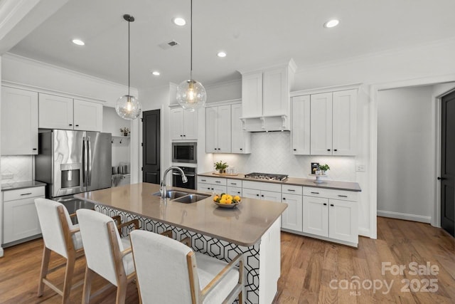 kitchen featuring a kitchen island with sink, ornamental molding, a sink, under cabinet range hood, and appliances with stainless steel finishes