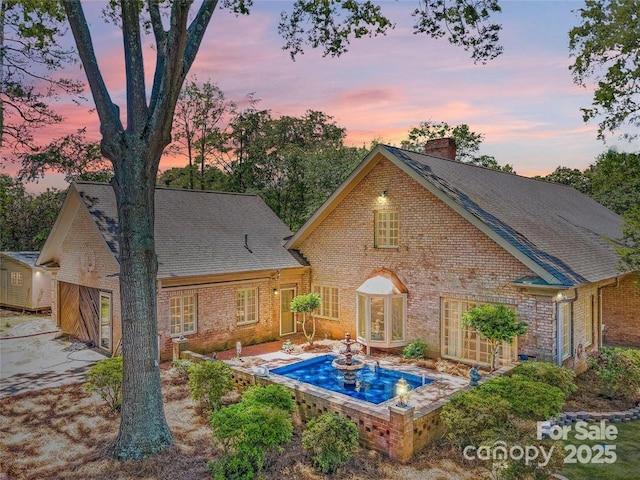 back of property at dusk featuring a shingled roof, a chimney, and brick siding