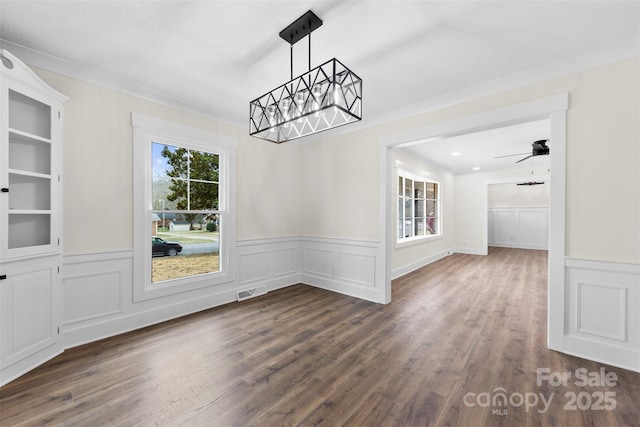 unfurnished dining area with dark wood-style floors, a wainscoted wall, ceiling fan with notable chandelier, and crown molding