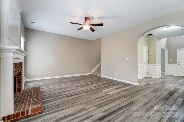 unfurnished living room featuring stairway, wood finished floors, arched walkways, ceiling fan, and a brick fireplace