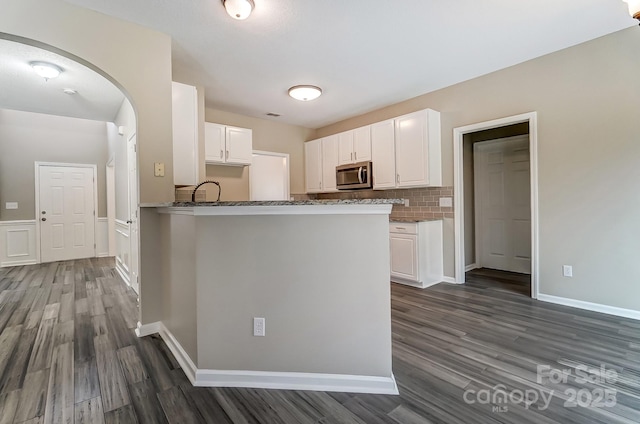 kitchen featuring stainless steel microwave, backsplash, dark stone counters, arched walkways, and white cabinetry