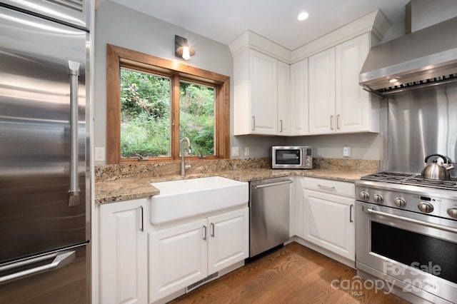 kitchen featuring high end appliances, wood finished floors, a sink, white cabinetry, and wall chimney range hood