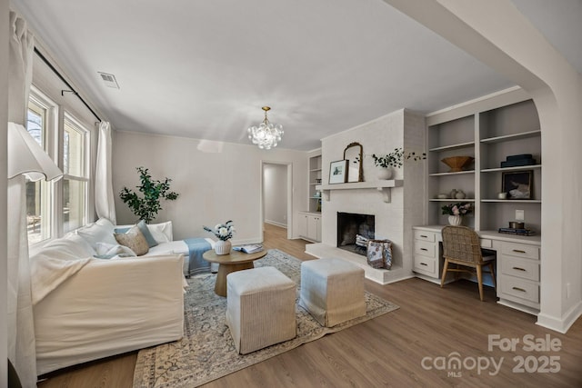 living room with built in shelves, visible vents, a brick fireplace, wood finished floors, and a chandelier