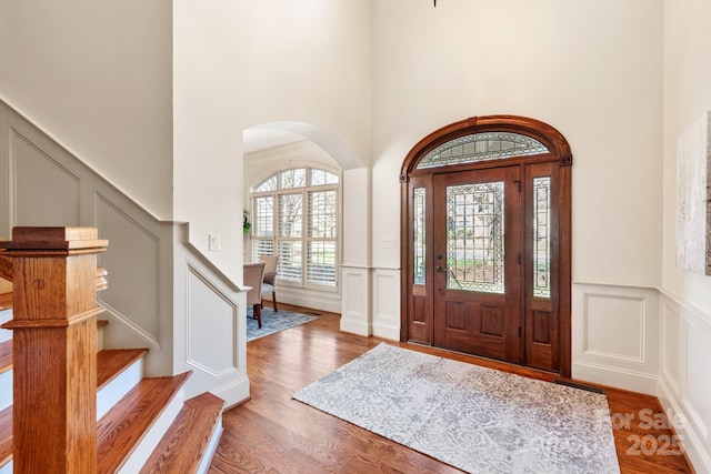 entrance foyer featuring arched walkways, a decorative wall, wood finished floors, stairs, and wainscoting