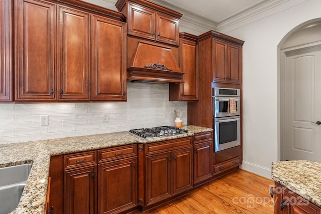 kitchen with light wood finished floors, tasteful backsplash, arched walkways, appliances with stainless steel finishes, and premium range hood