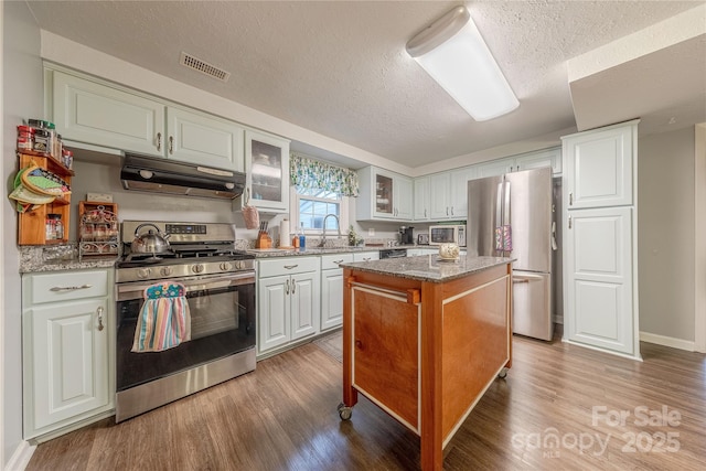 kitchen with under cabinet range hood, appliances with stainless steel finishes, glass insert cabinets, and wood finished floors