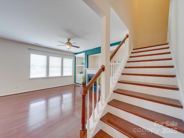 stairway with wood finished floors, visible vents, a ceiling fan, baseboards, and a glass covered fireplace