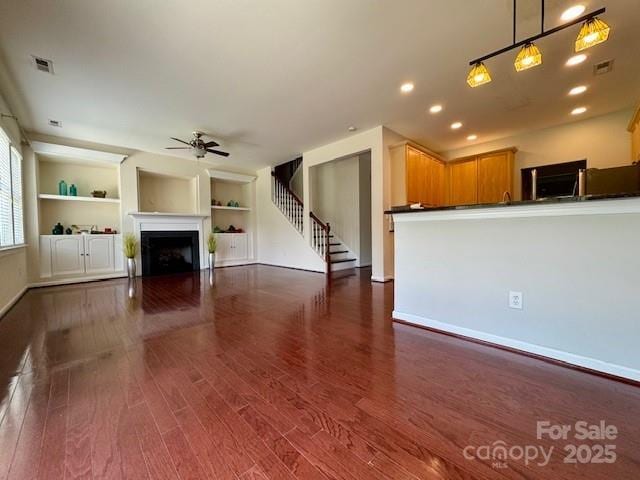 unfurnished living room with ceiling fan, dark wood-style floors, a fireplace, stairs, and built in shelves