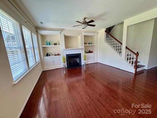 unfurnished living room featuring stairs, wood finished floors, ceiling fan, built in shelves, and a fireplace with flush hearth