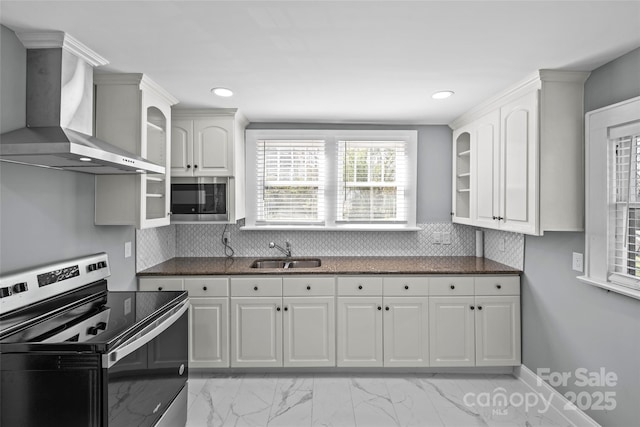 kitchen with appliances with stainless steel finishes, glass insert cabinets, white cabinetry, a sink, and wall chimney range hood