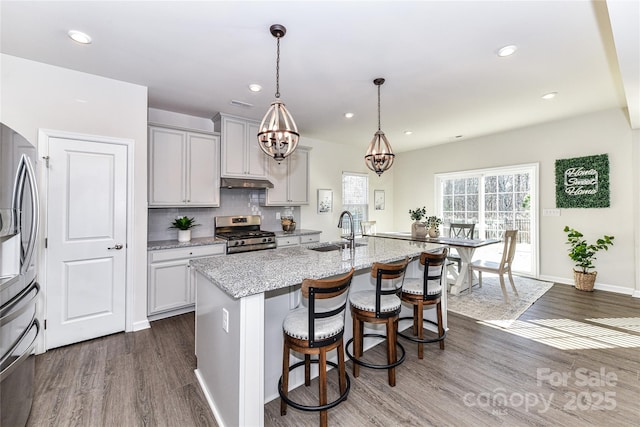 kitchen featuring a sink, under cabinet range hood, light stone counters, appliances with stainless steel finishes, and dark wood-style flooring