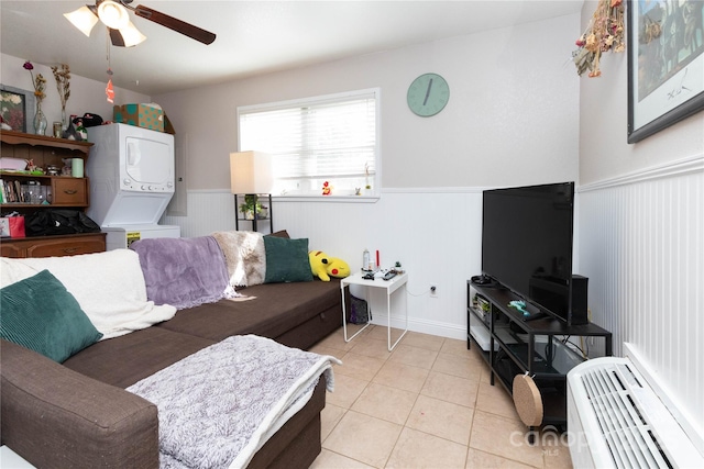 living area with a ceiling fan, light tile patterned flooring, stacked washer / dryer, and wainscoting