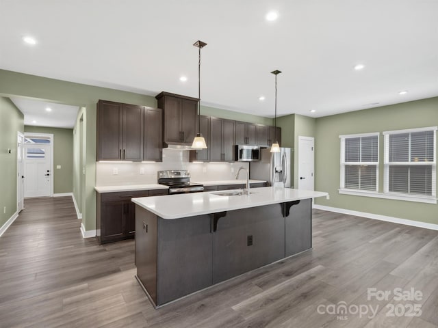 kitchen featuring a center island with sink, stainless steel appliances, light countertops, a sink, and wood finished floors