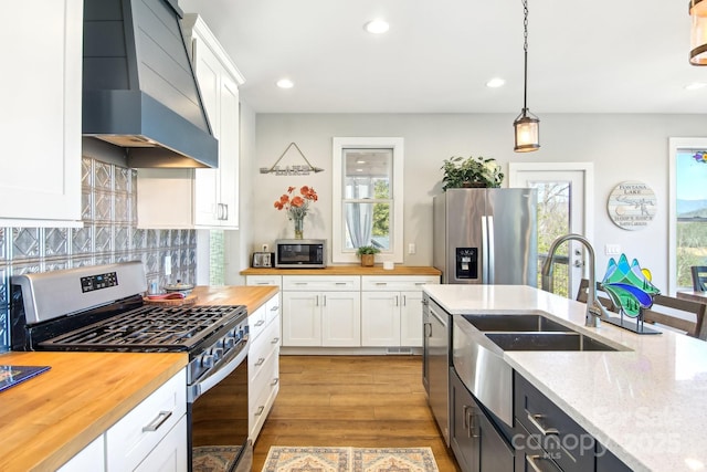 kitchen featuring wall chimney exhaust hood, butcher block countertops, stainless steel appliances, white cabinetry, and a sink