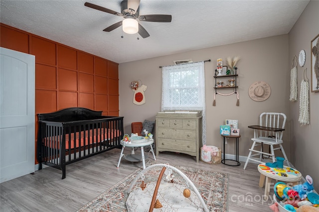 bedroom with light wood-type flooring, a nursery area, ceiling fan, and a textured ceiling