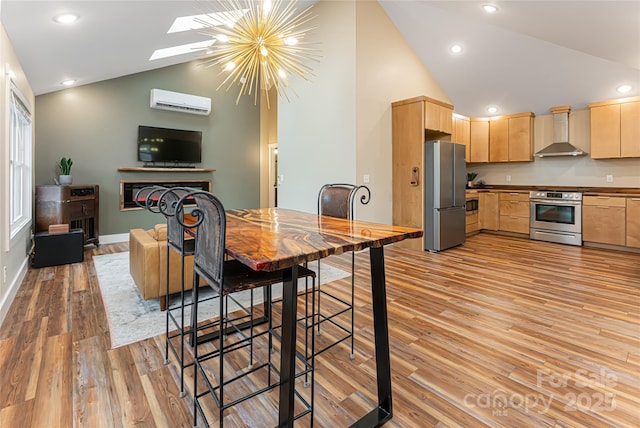 kitchen with stainless steel appliances, a warm lit fireplace, light brown cabinetry, and wall chimney range hood