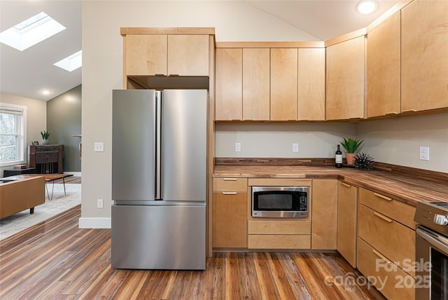 kitchen featuring dark wood finished floors, vaulted ceiling with skylight, light brown cabinetry, appliances with stainless steel finishes, and wood counters