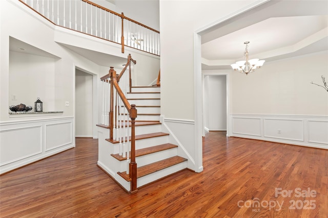 stairway with a raised ceiling, a decorative wall, wainscoting, wood finished floors, and a chandelier