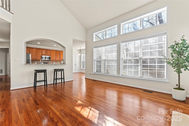 unfurnished living room with baseboards, visible vents, arched walkways, wood finished floors, and high vaulted ceiling