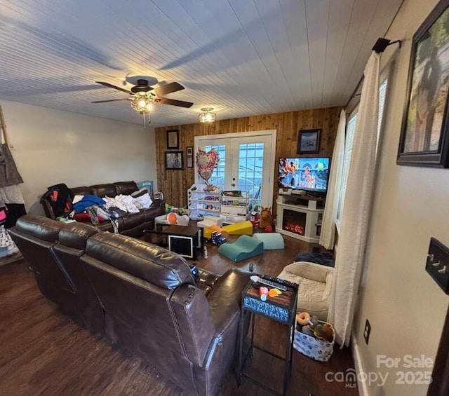 living area with french doors, ceiling fan, wooden walls, wood finished floors, and a lit fireplace