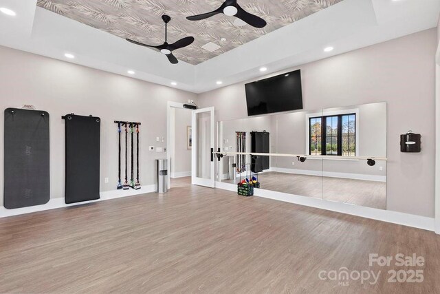 exercise room with a tray ceiling, wood finished floors, ceiling fan, and baseboards