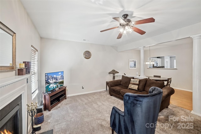 living room featuring decorative columns, a lit fireplace, baseboards, and ceiling fan with notable chandelier