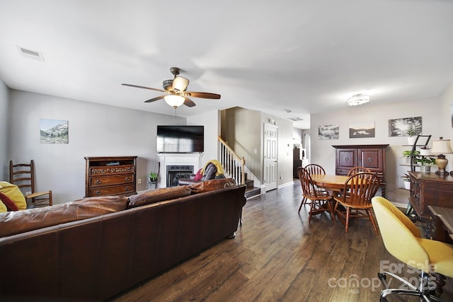 living area with visible vents, dark wood-type flooring, a fireplace, ceiling fan, and stairs