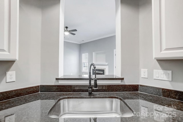 kitchen featuring a fireplace, ornamental molding, a sink, ceiling fan, and dark stone countertops