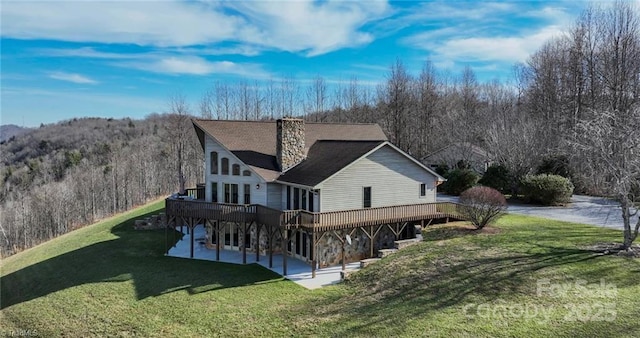 rear view of house featuring a view of trees, a chimney, a yard, a wooden deck, and a patio area