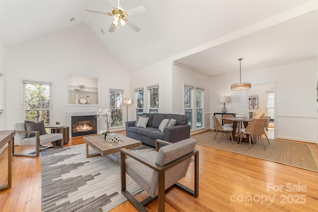 living area featuring high vaulted ceiling, light wood-type flooring, a glass covered fireplace, and a ceiling fan