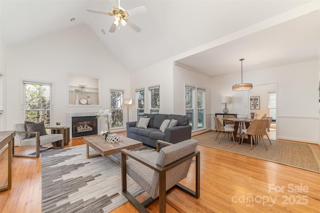 living room with high vaulted ceiling, a glass covered fireplace, light wood-style flooring, and a ceiling fan