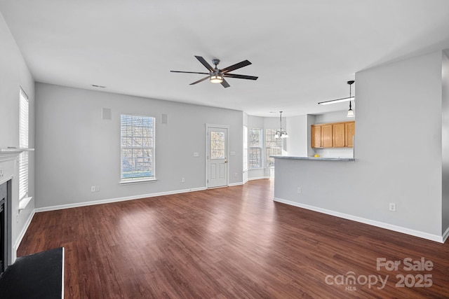 unfurnished living room featuring ceiling fan with notable chandelier, dark wood-style flooring, a fireplace, and a healthy amount of sunlight