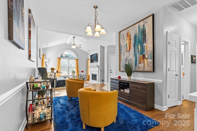 sitting room with a fireplace, visible vents, vaulted ceiling, wood finished floors, and ceiling fan with notable chandelier