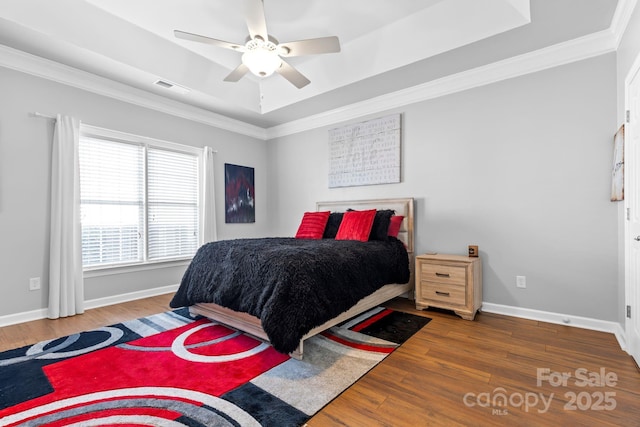 bedroom with wood finished floors, visible vents, baseboards, a raised ceiling, and crown molding