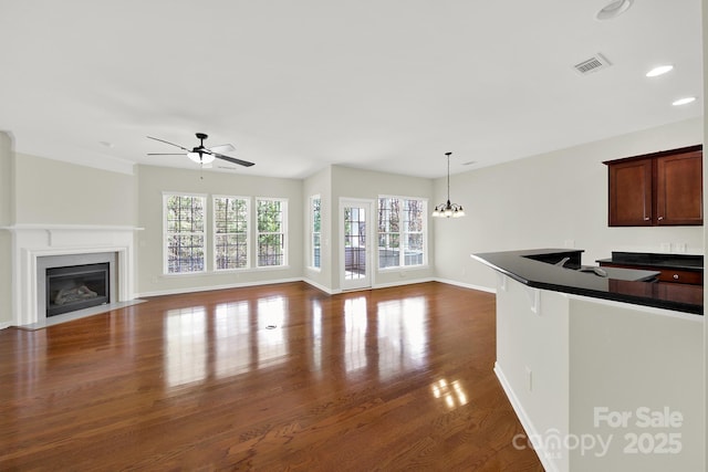 unfurnished living room with visible vents, baseboards, a fireplace with flush hearth, dark wood-type flooring, and ceiling fan with notable chandelier