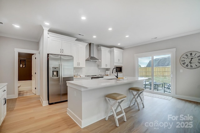 kitchen with appliances with stainless steel finishes, a sink, light wood finished floors, a kitchen island with sink, and wall chimney exhaust hood