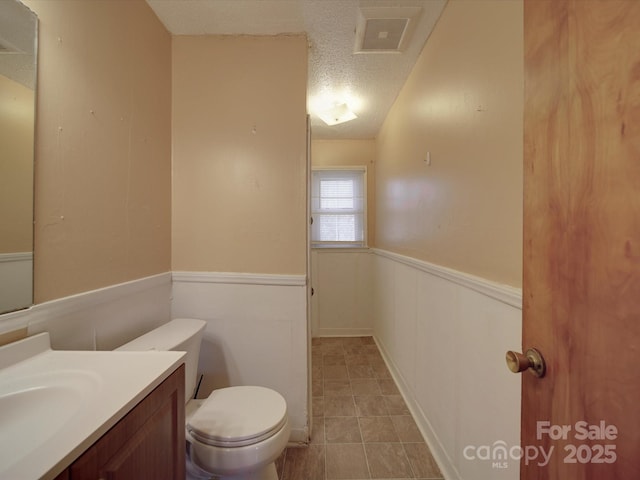 half bath with a wainscoted wall, visible vents, toilet, a textured ceiling, and vanity