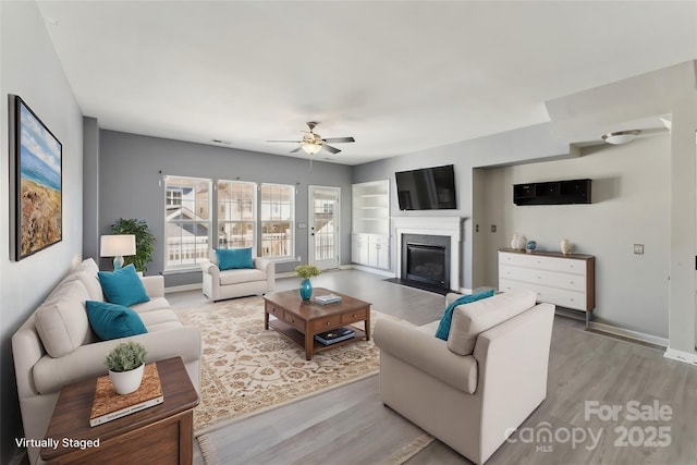 living room with light wood-style flooring, baseboards, a ceiling fan, and a fireplace with flush hearth