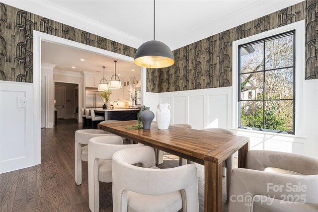 dining room with a wainscoted wall, wallpapered walls, dark wood-type flooring, crown molding, and a decorative wall