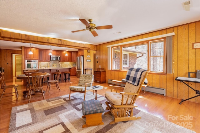 living room with wooden walls, a baseboard radiator, ceiling fan, a textured ceiling, and light wood-style floors