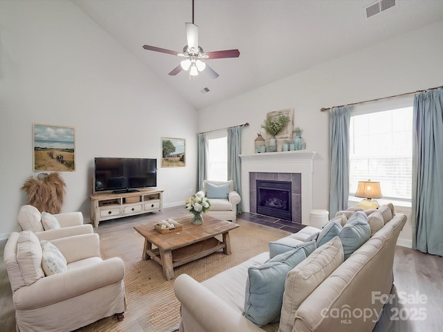 living area with ceiling fan, high vaulted ceiling, a tile fireplace, light wood-style flooring, and visible vents