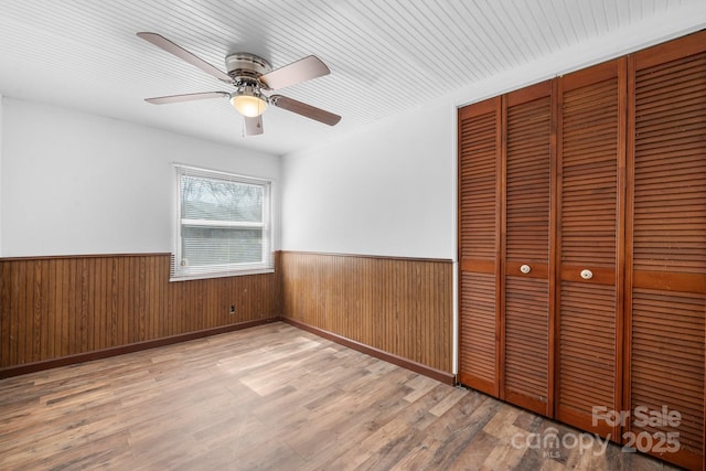 unfurnished bedroom featuring wooden walls, a ceiling fan, wood finished floors, a wainscoted wall, and a closet