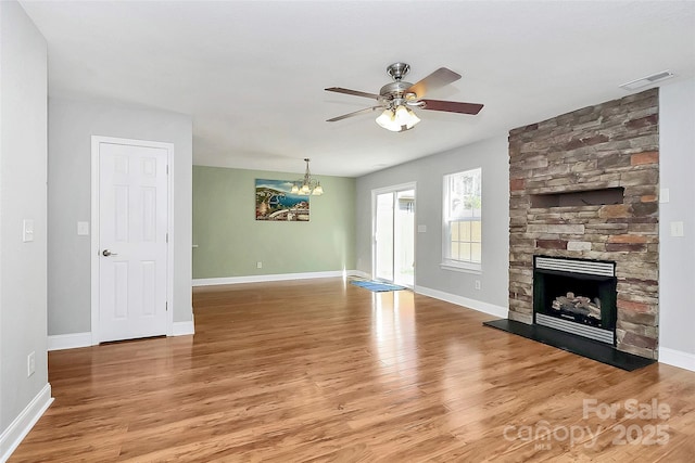 unfurnished living room with a stone fireplace, ceiling fan with notable chandelier, visible vents, baseboards, and light wood-style floors