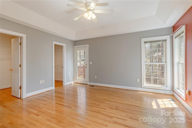 empty room with a ceiling fan, a tray ceiling, light wood-style floors, and baseboards