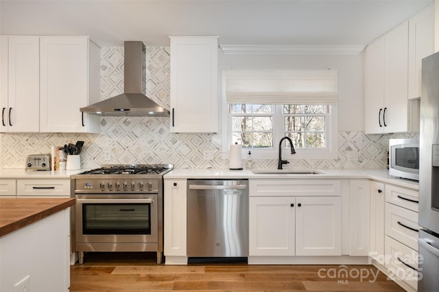 kitchen featuring wall chimney range hood, butcher block counters, ornamental molding, appliances with stainless steel finishes, and a sink