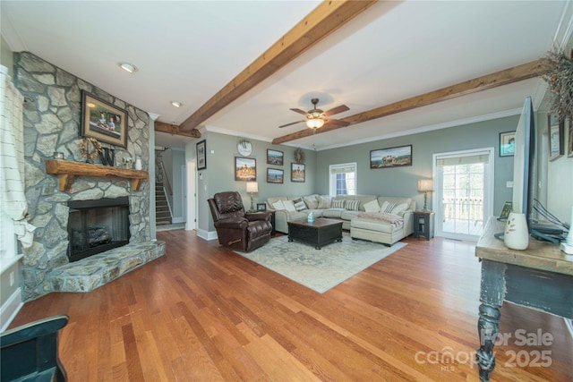 living room with a stone fireplace, ornamental molding, beamed ceiling, and wood finished floors