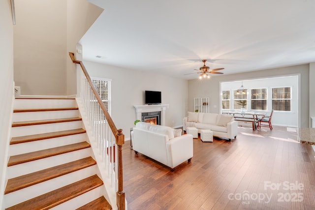 living area featuring a wealth of natural light, stairway, a lit fireplace, ceiling fan, and dark wood-style flooring