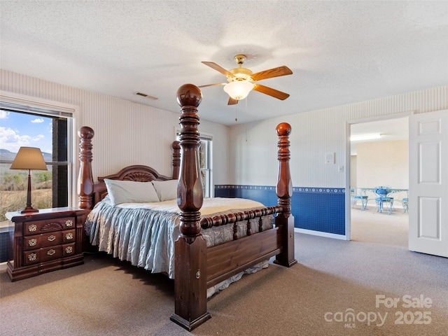 carpeted bedroom with visible vents, a textured ceiling, wainscoting, wallpapered walls, and ceiling fan