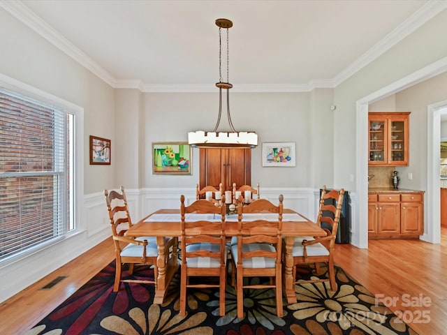 dining room with light wood-style floors, a wainscoted wall, visible vents, and crown molding