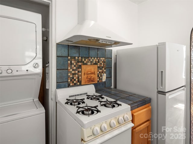 kitchen featuring white appliances, stacked washer and dryer, tile counters, decorative backsplash, and exhaust hood